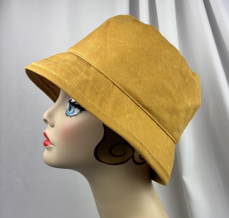 Puede incluir: Un sombrero de pescador color amarillo mostaza con copa redondeada y ala inclinada hacia abajo. El sombrero presenta costuras visibles a lo largo del ala y est&aacute; confeccionado con una tela de color s&oacute;lido con textura mate, sobre una cabeza de maniqu&iacute;.