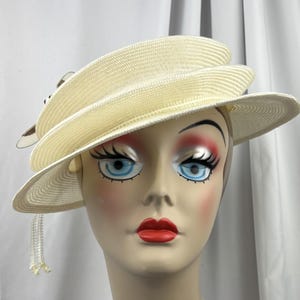 May include: A cream-colored straw hat with a multi-layered, asymmetrical brim design. The hat features a decorative accent on the side with small, dangling white beaded fringe elements that hang down from the edge of the brim.