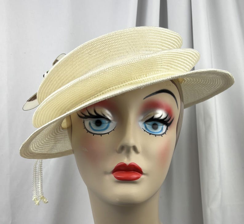 May include: A cream-colored straw hat with a multi-layered, asymmetrical brim design. The hat features a decorative accent on the side with small, dangling white beaded fringe elements that hang down from the edge of the brim.