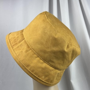 Puede incluir: Un sombrero de pescador color amarillo mostaza con una copa redondeada y un ala inclinada hacia abajo. El sombrero presenta costuras visibles a lo largo del ala y alrededor de la copa, confeccionado en tela de color s&oacute;lido con un acabado texturizado.