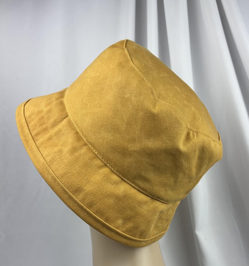 Puede incluir: Un sombrero de pescador color amarillo mostaza con una copa redondeada y un ala inclinada hacia abajo. El sombrero presenta costuras visibles a lo largo del ala y alrededor de la copa, confeccionado en tela de color s&oacute;lido con un acabado texturizado.