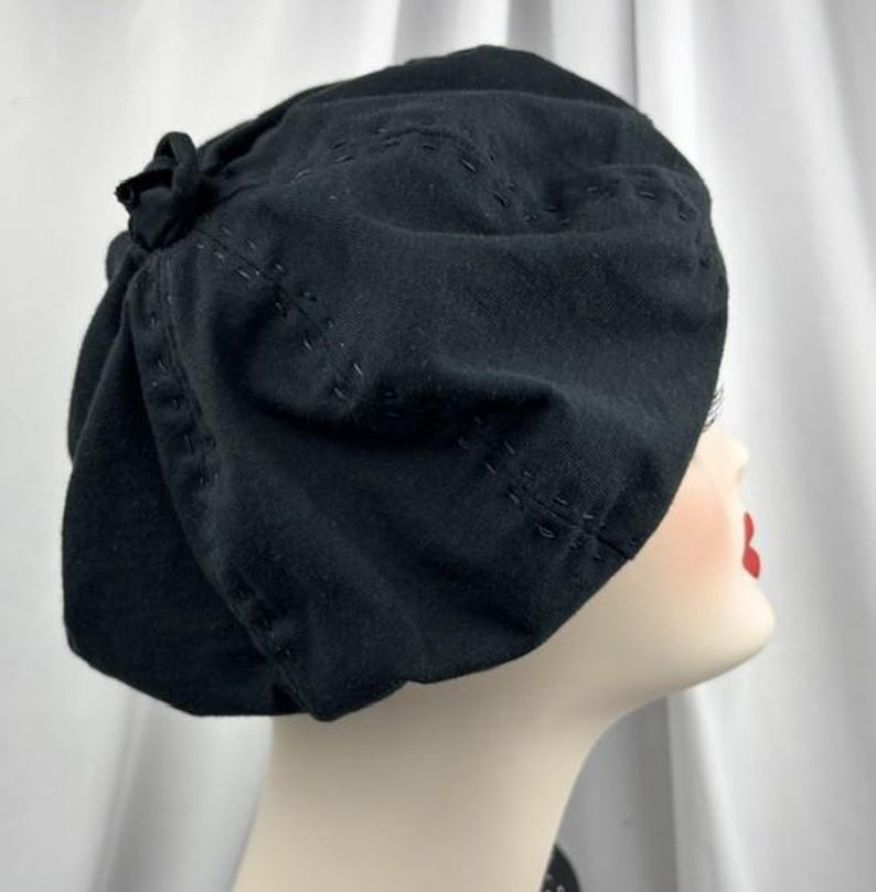 May include: A black beret featuring a soft, rounded crown and a small fabric knot detail at the top. The hat is constructed with visible, decorative tonal stitching that creates subtle, curved patterns across the fabric surface.