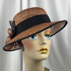 Pu&ograve; includere: Un cappello di paglia marrone con un nastro nero e un bordo nero. Il cappello ha un'ampia tesa e una piccola corona.