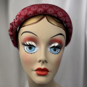 May include: A burgundy fabric headband with a repeating floral pattern and beaded embellishments. The headband is displayed on a mannequin head with painted features, including blue eyes and red lips. The headband is a fashion accessory.