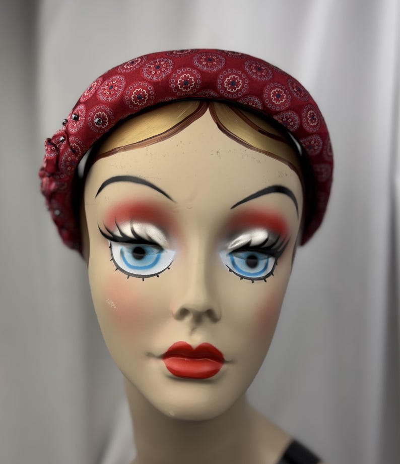 May include: A burgundy fabric headband with a repeating floral pattern and beaded embellishments. The headband is displayed on a mannequin head with painted features, including blue eyes and red lips. The headband is a fashion accessory.