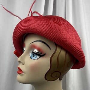 May include: A bright red straw hat with a rounded crown and a curved brim. The hat is adorned with thin, delicate red feathers extending from the side, adding a textured detail to the solid red woven material of the hat's exterior structure.