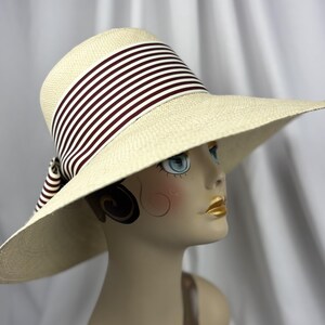 Pu&ograve; includere: Un cappello di paglia a tesa larga con corona e tesa color crema. Il cappello presenta un nastro a righe bordeaux e bianco attorno alla corona. Il cappello &egrave; esposto su una testa di manichino.