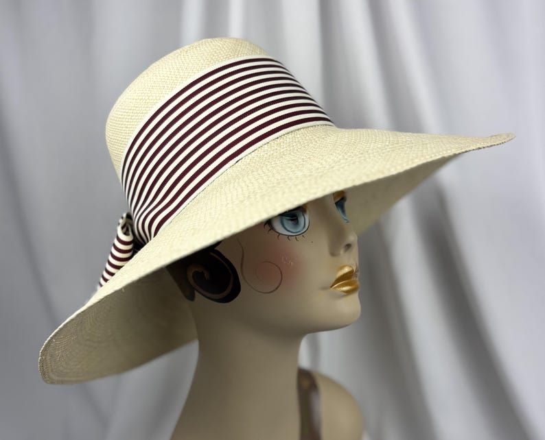 Pu&ograve; includere: Un cappello di paglia a tesa larga con corona e tesa color crema. Il cappello presenta un nastro a righe bordeaux e bianco attorno alla corona. Il cappello &egrave; esposto su una testa di manichino.