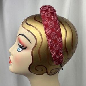 May include: A red fabric headband with a repeating circular pattern in white, blue, and red. The headband is on a mannequin head with gold-painted hair and facial features. A small fabric flower is attached to the side.
