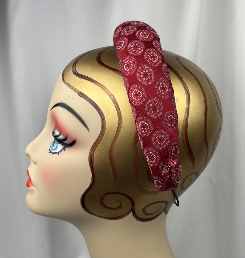 May include: A red fabric headband with a repeating circular pattern in white, blue, and red. The headband is on a mannequin head with gold-painted hair and facial features. A small fabric flower is attached to the side.