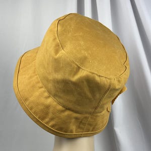 Puede incluir: Un sombrero de pescador color amarillo mostaza con una copa redondeada y un ala inclinada hacia abajo. El sombrero presenta costuras visibles a lo largo del ala y la copa, con una superficie de tela texturizada que tiene un aspecto desgastado.