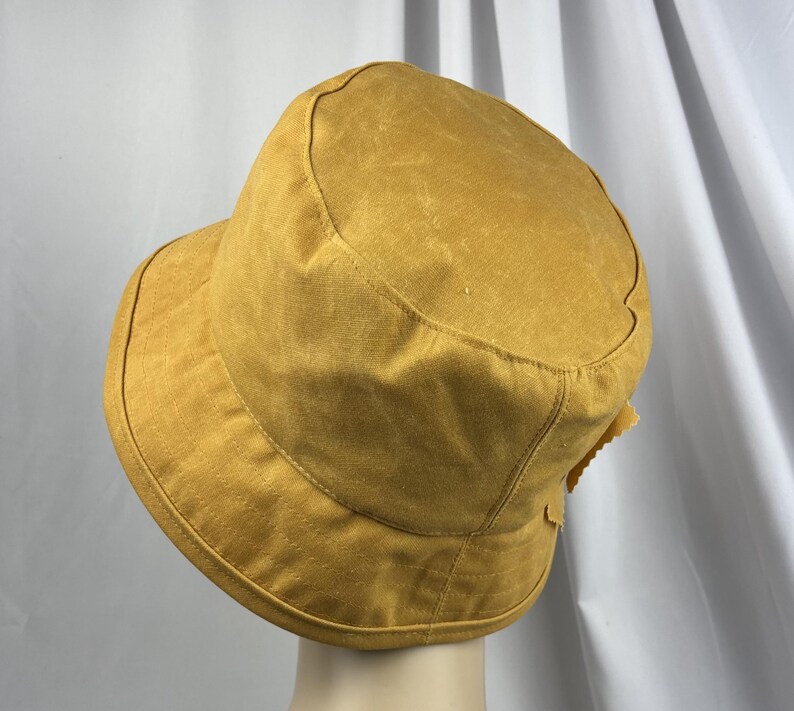 Puede incluir: Un sombrero de pescador color amarillo mostaza con una copa redondeada y un ala inclinada hacia abajo. El sombrero presenta costuras visibles a lo largo del ala y la copa, con una superficie de tela texturizada que tiene un aspecto desgastado.