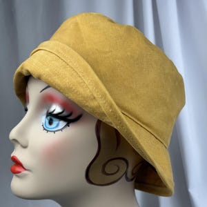 Puede incluir: Un sombrero de pescador de color amarillo mostaza con un ala ancha inclinada hacia abajo y una copa redondeada. El sombrero est&aacute; confeccionado con un tejido texturizado y presenta costuras visibles a lo largo del ala, con forma firme.