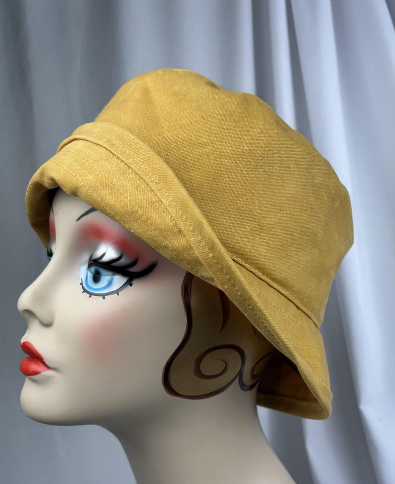 Puede incluir: Un sombrero de pescador de color amarillo mostaza con un ala ancha inclinada hacia abajo y una copa redondeada. El sombrero est&aacute; confeccionado con un tejido texturizado y presenta costuras visibles a lo largo del ala, con forma firme.