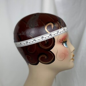 May include: A mannequin head with painted brown hair in a finger wave style, blue eyes, and red lips. A white measuring tape wraps around the head, showing a circumference measurement of approximately 18 inches at the widest point of the crown.