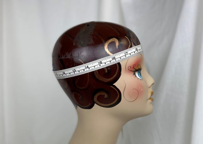 May include: A mannequin head with painted brown hair in a finger wave style, blue eyes, and red lips. A white measuring tape wraps around the head, showing a circumference measurement of approximately 18 inches at the widest point of the crown.