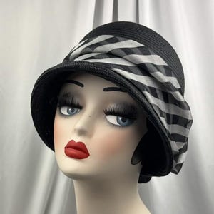 May include: Black cloche hat with a wide brim and a striped ribbon detail. The hat is made of woven material and features a contrasting black and white pattern on the ribbon. The hat is displayed on a mannequin head.