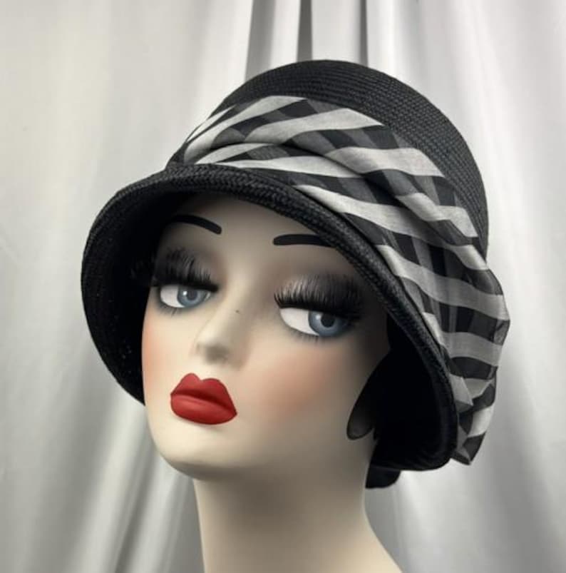 May include: Black cloche hat with a wide brim and a striped ribbon detail. The hat is made of woven material and features a contrasting black and white pattern on the ribbon. The hat is displayed on a mannequin head.