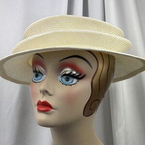 May include: A cream-colored straw hat featuring a multi-tiered, layered crown design and a wide, structured brim. The hat is constructed from a tightly woven material with a consistent texture throughout, showcasing a clean, circular silhouette.