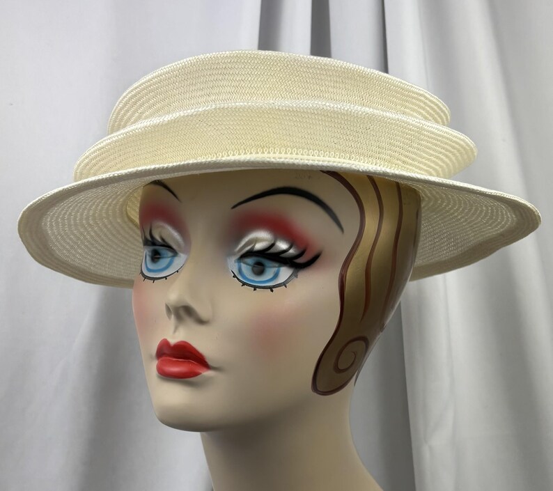 May include: A cream-colored straw hat featuring a multi-tiered, layered crown design and a wide, structured brim. The hat is constructed from a tightly woven material with a consistent texture throughout, showcasing a clean, circular silhouette.