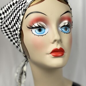 May include: A mannequin head wearing a black and white checkered headscarf. The headscarf is tied at the back. The mannequin has blue eyes, red lips, and blush. The headscarf is a fashion accessory.