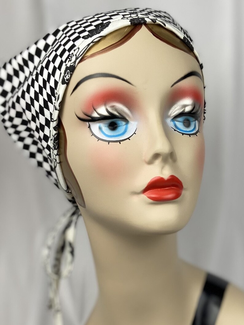 May include: A mannequin head wearing a black and white checkered headscarf. The headscarf is tied at the back. The mannequin has blue eyes, red lips, and blush. The headscarf is a fashion accessory.