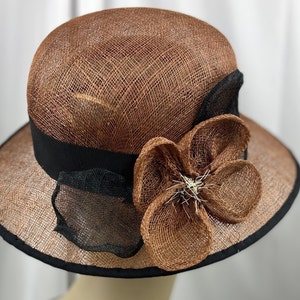 Pu&ograve; includere: Un cappello di paglia marrone con un nastro nero e un grande fiore marrone fatto di paglia.