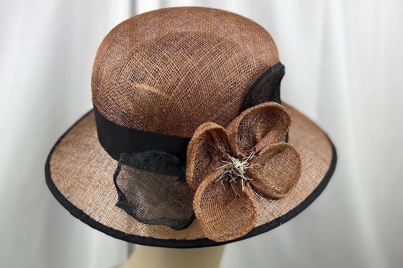 Pu&ograve; includere: Un cappello di paglia marrone con un nastro nero e un grande fiore marrone fatto di paglia.