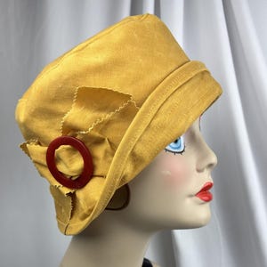 Puede incluir: Un sombrero tipo cloche de color amarillo mostaza con una copa estructurada y un ala inclinada hacia abajo. El lateral del sombrero est&aacute; adornado con un lazo de tela y un anillo circular de pl&aacute;stico rojo oscuro, creando un contraste.