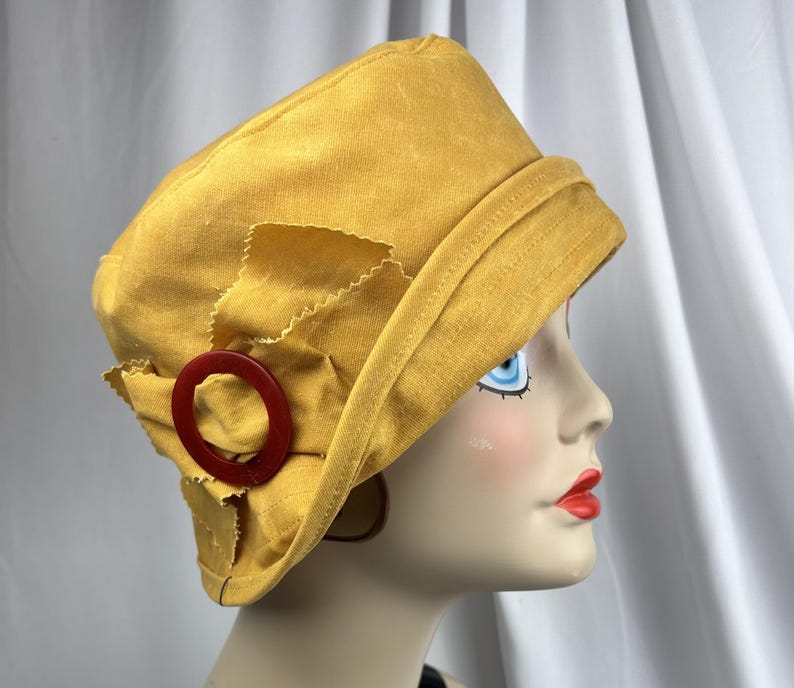Puede incluir: Un sombrero tipo cloche de color amarillo mostaza con una copa estructurada y un ala inclinada hacia abajo. El lateral del sombrero est&aacute; adornado con un lazo de tela y un anillo circular de pl&aacute;stico rojo oscuro, creando un contraste.