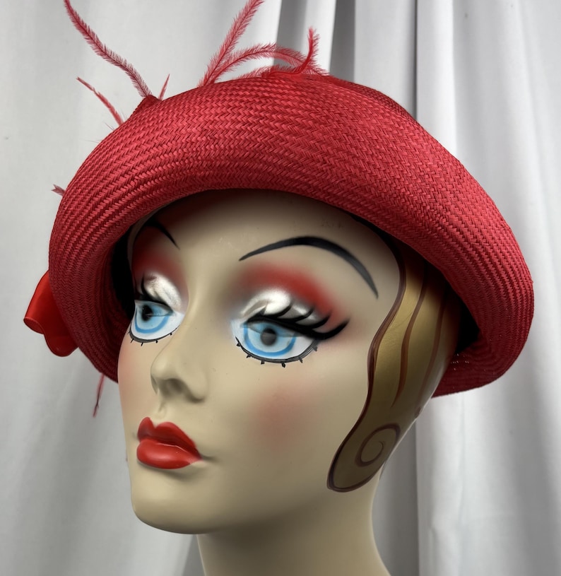 May include: A bright red woven straw hat featuring a wide, structured brim and a decorative red ribbon bow on the side. The hat is adorned with thin, delicate red feathers that extend upwards from the crown, adding texture to the design.