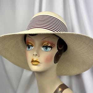 Pu&ograve; includere: Un cappello da sole color crema con tesa larga e fascia a righe bianche e bordeaux. Il cappello &egrave; esposto su una testa di manichino. L'ampia tesa offre protezione solare. La fascia ha strisce bianche e bordeaux.
