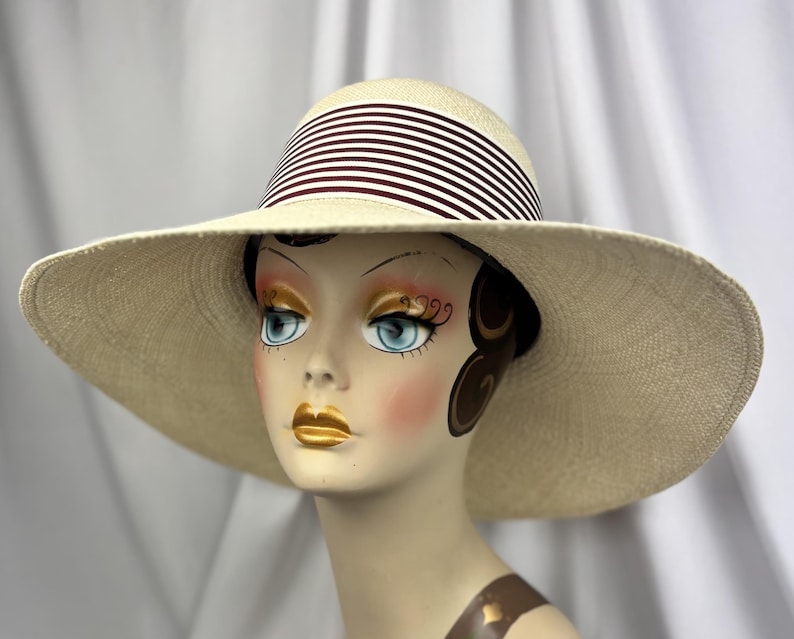 Pu&ograve; includere: Un cappello da sole color crema con tesa larga e fascia a righe bianche e bordeaux. Il cappello &egrave; esposto su una testa di manichino. L'ampia tesa offre protezione solare. La fascia ha strisce bianche e bordeaux.