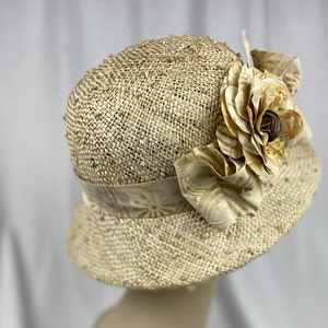 Puede incluir: Un sombrero de paja de ala ancha con una gran flor de tela en tonos beige y marr&oacute;n.