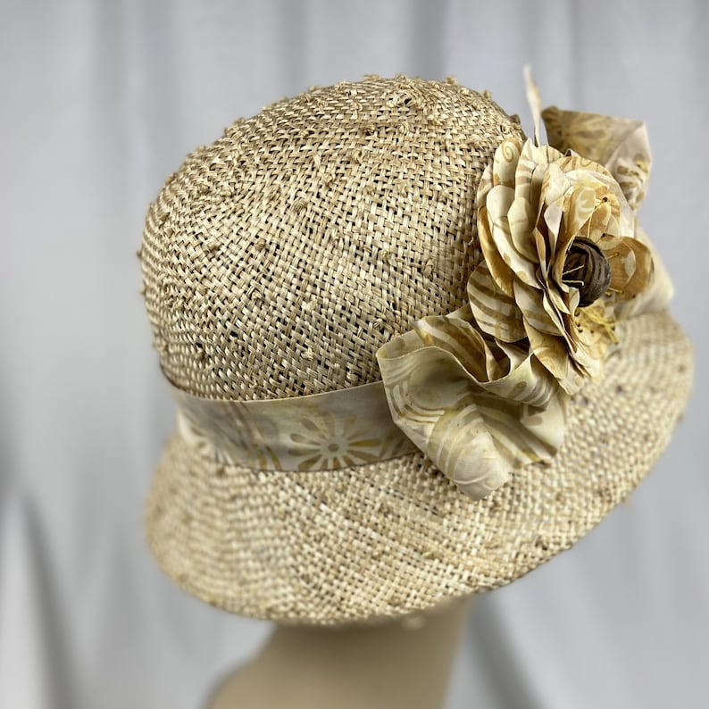 Puede incluir: Un sombrero de paja de ala ancha con una gran flor de tela en tonos beige y marr&oacute;n.