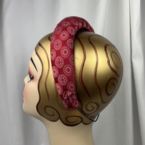 May include: A padded headband with a burgundy fabric featuring a repeating circular pattern in red, white, and blue. The headband is displayed on a mannequin head with gold paint and brown swirls.