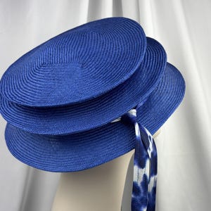 K&ouml;nnte beinhalten: Ein leuchtend k&ouml;nigsblauer Hut mit einer mehrstufigen, kreisf&ouml;rmigen stroh&auml;hnlichen Konstruktion und einem markanten Schichtdesign. Ein langes, flie&szlig;endes Band mit wei&szlig;-blauem Batikmuster ist seitlich befestigt und h&auml;ngt vom Rand herab.