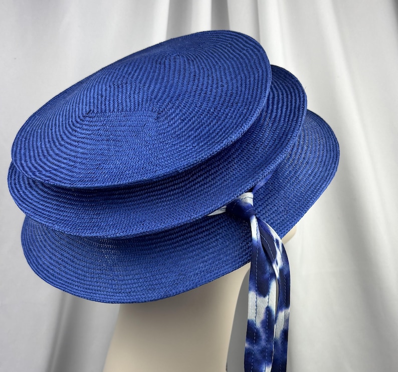 K&ouml;nnte beinhalten: Ein leuchtend k&ouml;nigsblauer Hut mit einer mehrstufigen, kreisf&ouml;rmigen stroh&auml;hnlichen Konstruktion und einem markanten Schichtdesign. Ein langes, flie&szlig;endes Band mit wei&szlig;-blauem Batikmuster ist seitlich befestigt und h&auml;ngt vom Rand herab.