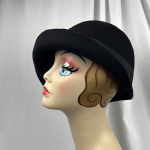 May include: A solid black felt cloche hat with a rounded crown and a slightly downturned brim. The hat features a simple black ribbon band encircling the base of the crown, providing a clean and uniform appearance on the mannequin head.
