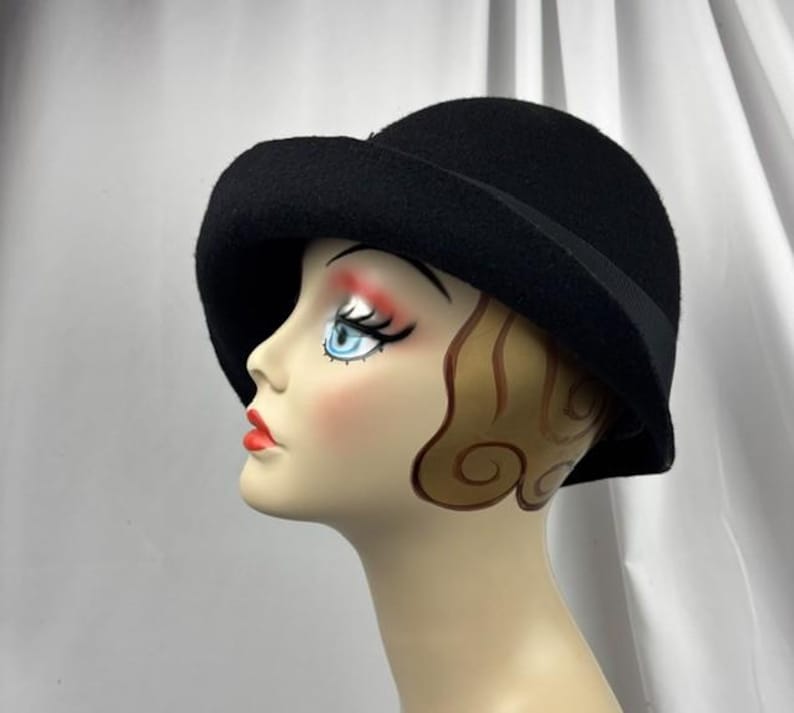 May include: A solid black felt cloche hat with a rounded crown and a slightly downturned brim. The hat features a simple black ribbon band encircling the base of the crown, providing a clean and uniform appearance on the mannequin head.