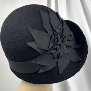 May include: A black felt cloche hat featuring a wide grosgrain ribbon band around the base and a large, layered fabric flower ornament on the side. The flower is constructed from multiple pointed, folded black ribbon petals.