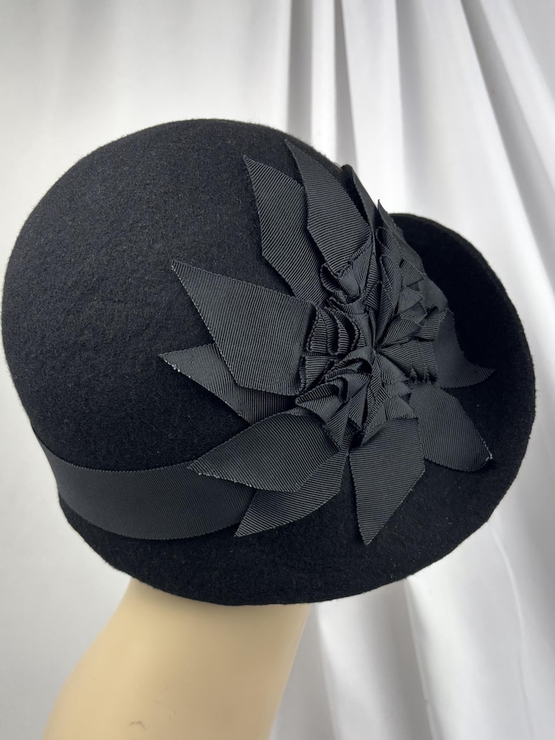 May include: A black felt cloche hat featuring a wide grosgrain ribbon band around the base and a large, layered fabric flower ornament on the side. The flower is constructed from multiple pointed, folded black ribbon petals.