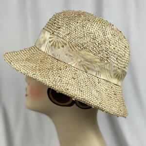 Puede incluir: Un sombrero de paja de ala ancha con una cinta de estampado marr&oacute;n. El sombrero est&aacute; hecho de un material tejido y tiene una superficie ligeramente texturizada.