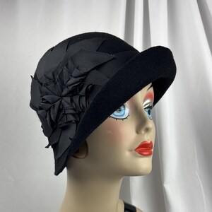May include: A black felt cloche hat featuring a wide, asymmetrical brim and a decorative black ribbon flower accent on the side. The hat is constructed from a solid black material with a textured finish and a structured, rounded crown shape.