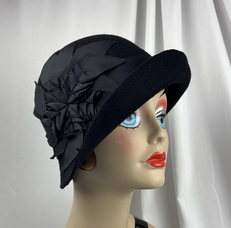 May include: A black felt cloche hat featuring a wide, asymmetrical brim and a decorative black ribbon flower accent on the side. The hat is constructed from a solid black material with a textured finish and a structured, rounded crown shape.