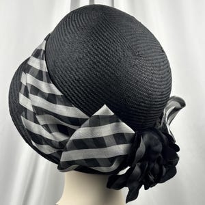 May include: A black hat with a rounded crown and a wide brim. The hat features a black and white checked ribbon and a black fabric flower. The hat is made of woven material.