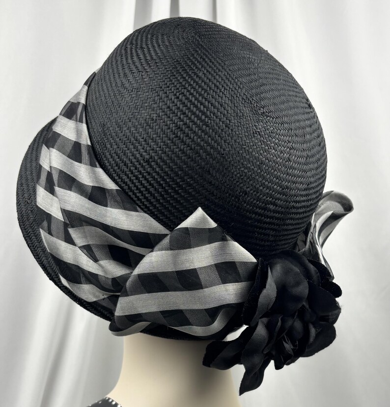 May include: A black hat with a rounded crown and a wide brim. The hat features a black and white checked ribbon and a black fabric flower. The hat is made of woven material.