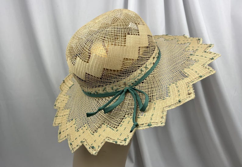 May include: A woven straw hat with a wide, scalloped brim. The hat features a geometric pattern on the crown and a green ribbon tied in a bow. The brim is trimmed with green stitching. The hat is a light tan color.