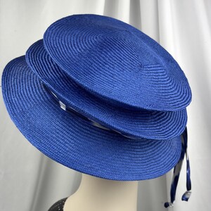 K&ouml;nnte beinhalten: Ein leuchtend k&ouml;nigsblauer Hut aus eng geflochtenem Strohmaterial. Der Hut zeichnet sich durch ein markantes, gestuftes Design mit drei konzentrischen, kreisf&ouml;rmigen Lagen unterschiedlicher Gr&ouml;&szlig;e aus, die vertikal &uuml;bereinander liegen.
