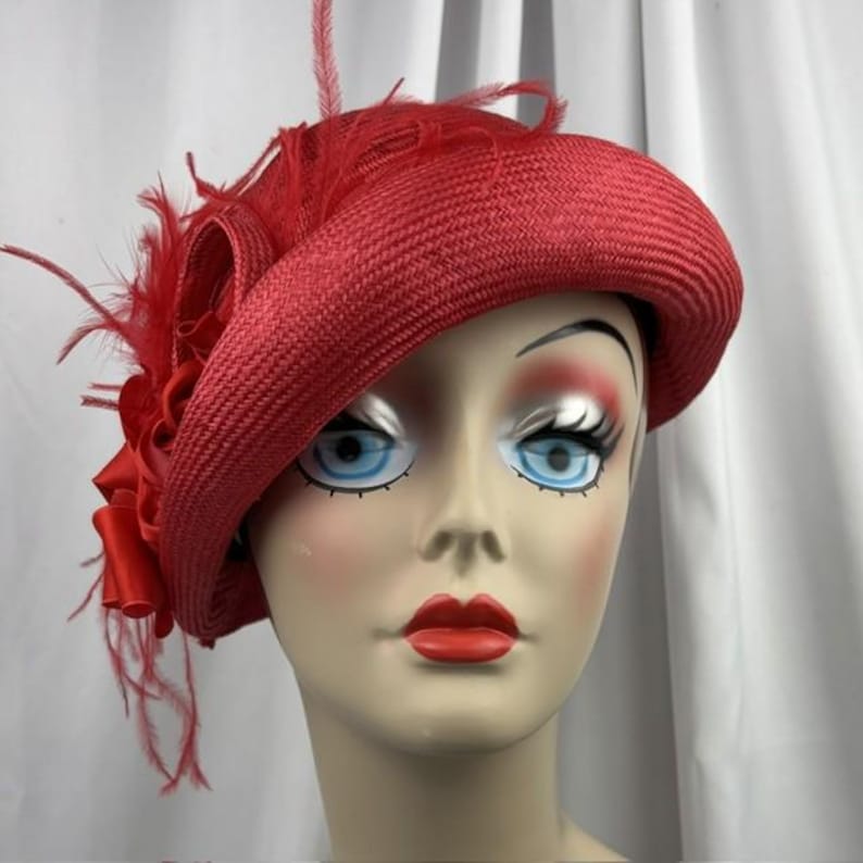 May include: A vibrant red straw hat featuring a wide, structured brim and a decorative side accent composed of matching red satin ribbon loops and delicate, wispy feathers that extend upward and outward from the side of the crown.